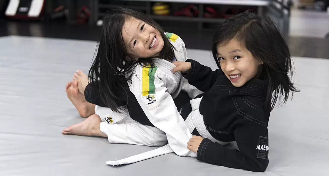 Two young girls in jiu-jitsu gis laughing as they grapple on the mats at Trillium MMA